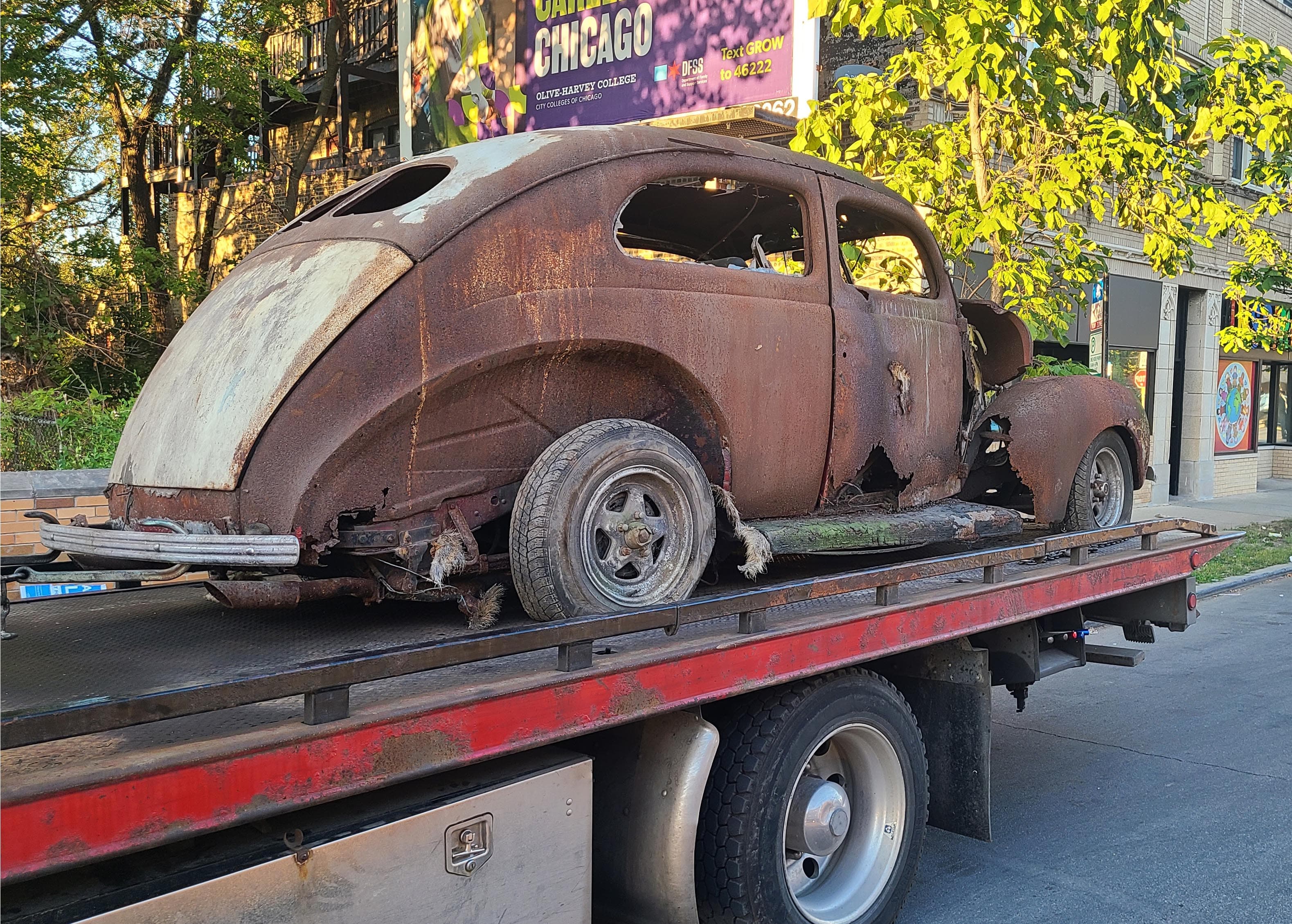 Heavily rusted vintage car with missing windows on a flatbed tow truck in a city.