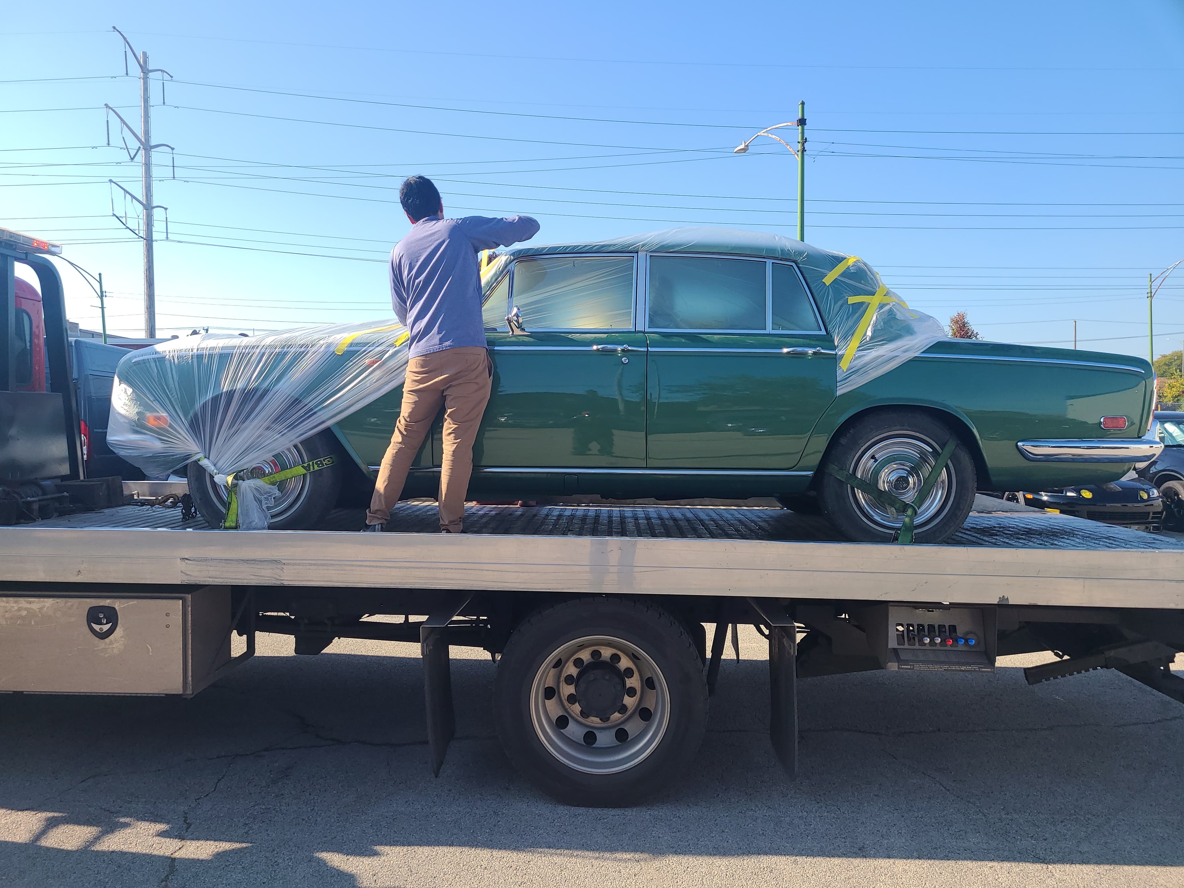 Man wraps a green vintage car in protective plastic on a flatbed tow truck.